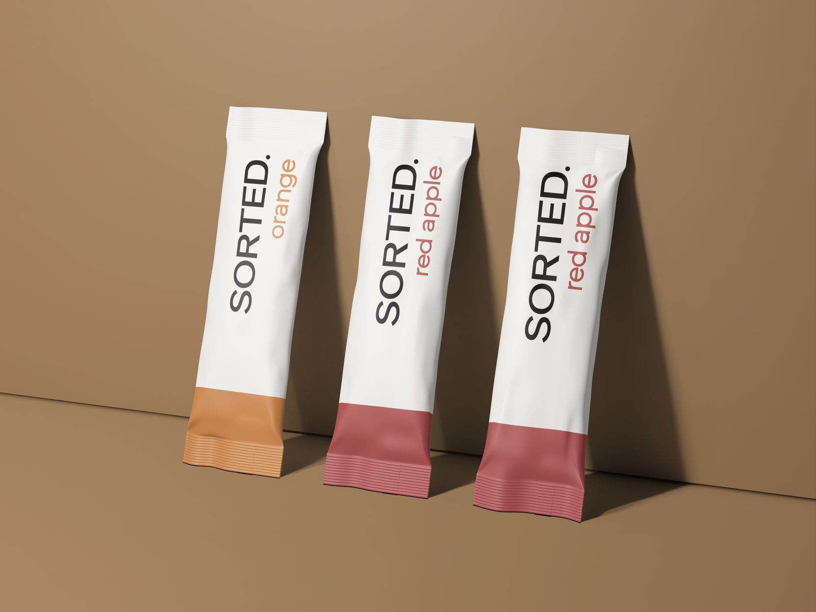 Three SORTED. single-serve sachets, one orange and two red apple, standing against a tan wall